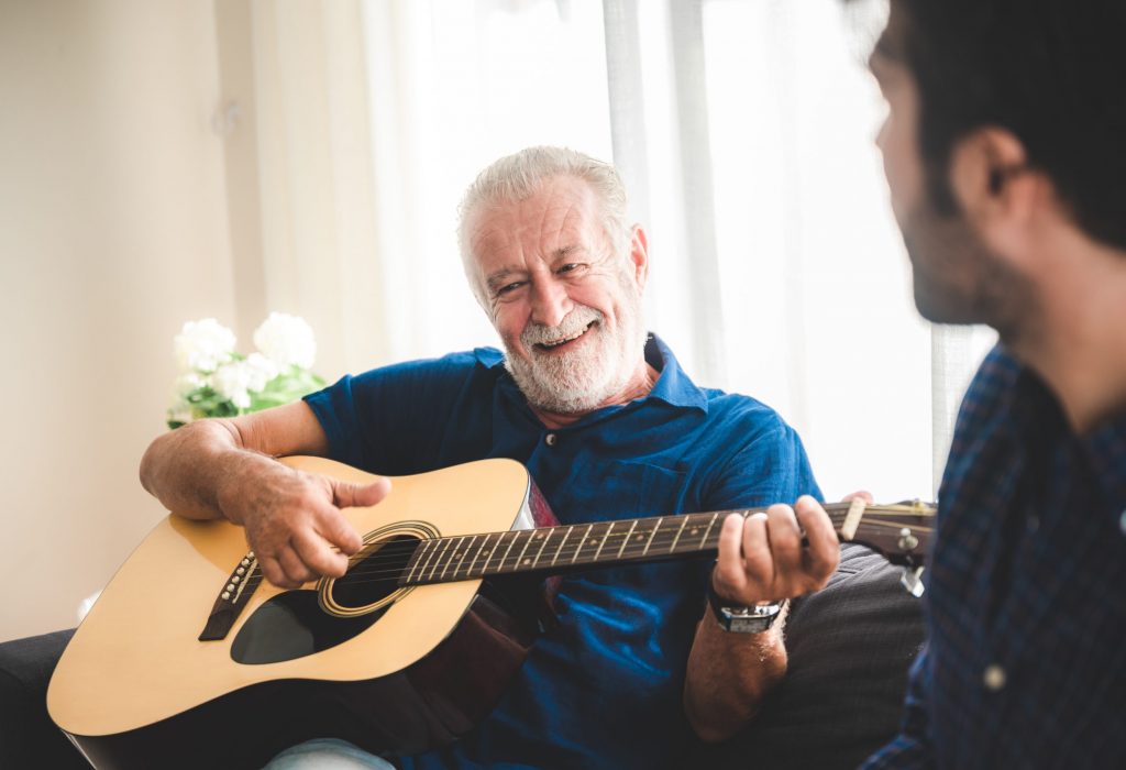 happy adult son and senior father playing guitar on sofa at home relaxing and having happy time with family